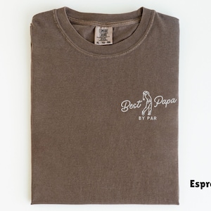 Op de afbeelding: Espresso-kleurig T-shirt met de witte tekst "Best Papa by Par". Het shirt heeft een afbeelding van een golfer. Het shirt is opgevouwen en heeft een label in de linkerbovenhoek.