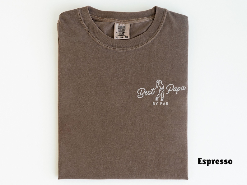 Op de afbeelding: Espresso-kleurig T-shirt met de witte tekst "Best Papa by Par". Het shirt heeft een afbeelding van een golfer. Het shirt is opgevouwen en heeft een label in de linkerbovenhoek.