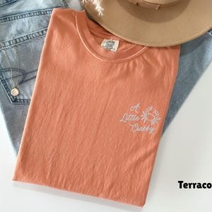 May include: Terracotta-colored t-shirt with the embroidered phrase "A Little Crabby" and a crab graphic. The shirt is laid on top of blue jeans and a tan hat.