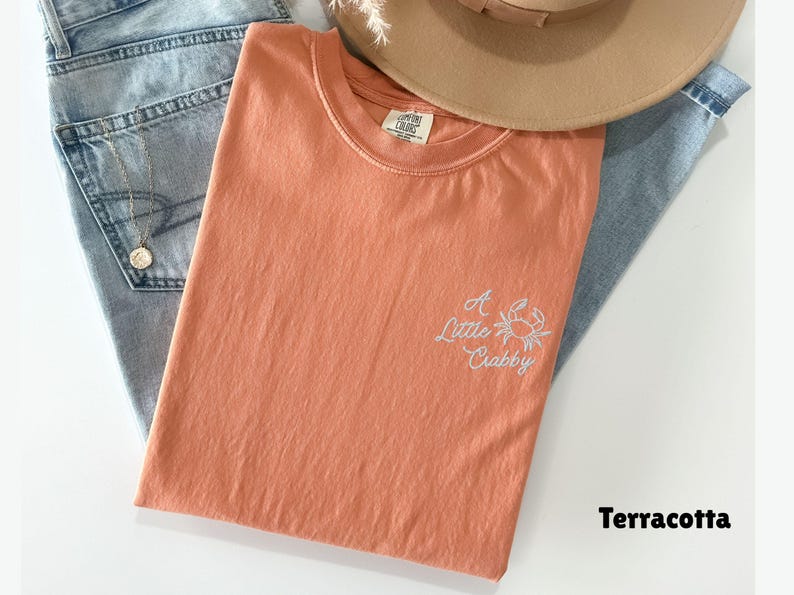 May include: Terracotta-colored t-shirt with the embroidered phrase "A Little Crabby" and a crab graphic. The shirt is laid on top of blue jeans and a tan hat.