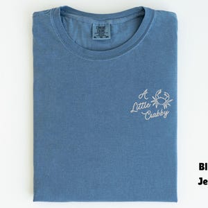 May include: Blue jean-colored t-shirt with a crew neck, folded neatly. The shirt features the embroidered phrase "A Little Crabby" with a crab graphic in white thread on the front. The shirt is a size XL.