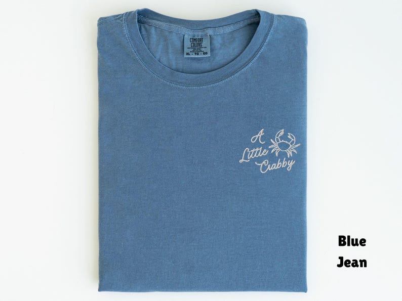 May include: Blue jean-colored t-shirt with a crew neck, folded neatly. The shirt features the embroidered phrase "A Little Crabby" with a crab graphic in white thread on the front. The shirt is a size XL.