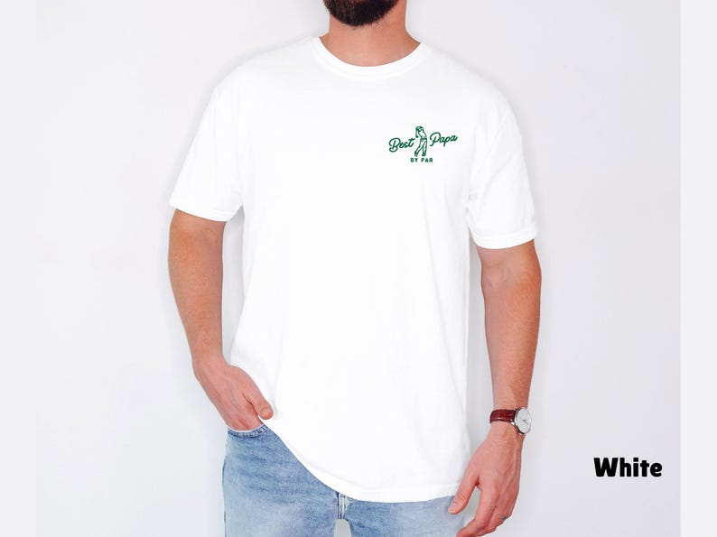 Op de afbeelding: Wit T-shirt met de tekst "Best Papa by Far" in groen schrift. Het shirt heeft een ronde hals. De persoon die het shirt draagt, draagt een blauwe spijkerbroek en een horloge met een bruine leren band.