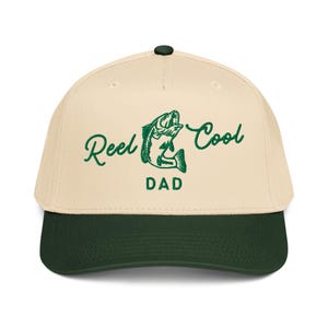 May include: A beige and dark green baseball cap with the words "Reel Cool Dad" in green script. The design includes a green fish graphic. The cap has a curved brim and a button on top.