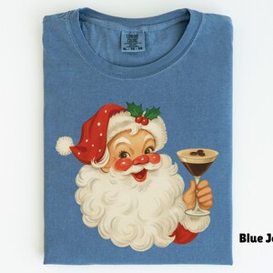 May include: Blue jean colored t-shirt featuring a vintage illustration of Santa Claus holding a cocktail. Santa is wearing a red hat with white polka dots and a white beard. The shirt has a classic crew neck design.