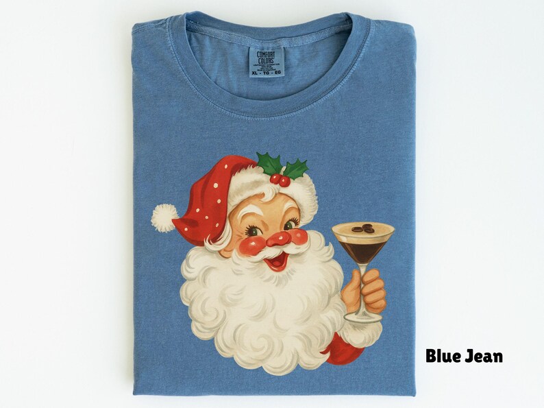 May include: Blue jean colored t-shirt featuring a vintage illustration of Santa Claus holding a cocktail. Santa is wearing a red hat with white polka dots and a white beard. The shirt has a classic crew neck design.