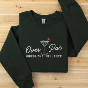 May include: Dark green sweatshirt with the text "Over Par Under the Influence" in white script. A martini glass with a red flag is in the center. The sweatshirt is folded on a wooden surface.