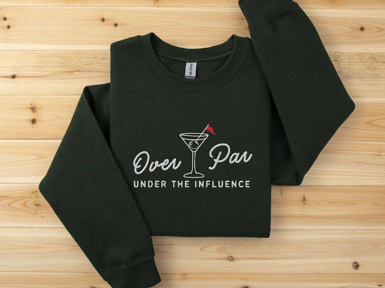 May include: Dark green sweatshirt with the text "Over Par Under the Influence" in white script. A martini glass with a red flag is in the center. The sweatshirt is folded on a wooden surface.