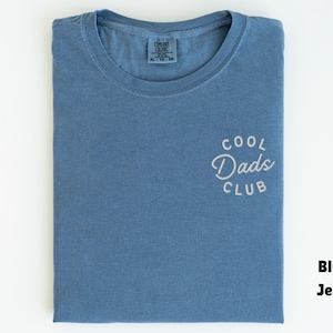 May include: A blue jean colored t-shirt with the text "COOL Dads CLUB" embroidered on the front. The shirt is folded neatly and has a crew neck. The shirt is a size XL.