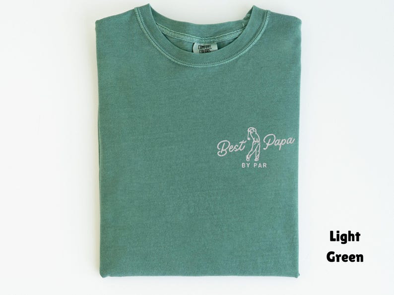 Op de afbeelding: Lichtgroen T-shirt met de tekst "Best Papa" en een golfafbeelding. Het shirt is opgevouwen en heeft een ronde hals. De woorden "BY PAR" staan onder de hoofdtekst. De woorden "Light Green" staan in de rechter benedenhoek.