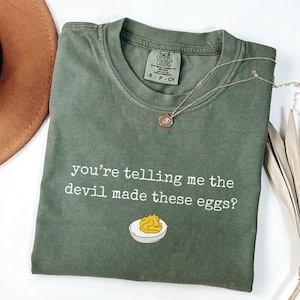 May include: Olive green t-shirt with the text "you're telling me the devil made these eggs?" and an illustration of a deviled egg. A gold necklace and a brown hat are also visible.