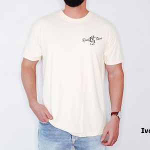 Pu&ograve; includere: T-shirt color avorio con la scritta "Reel Cool Dad" in corsivo nero. La maglietta ha girocollo e maniche corte. Il modello indossa jeans blu e un orologio con cinturino in pelle marrone.