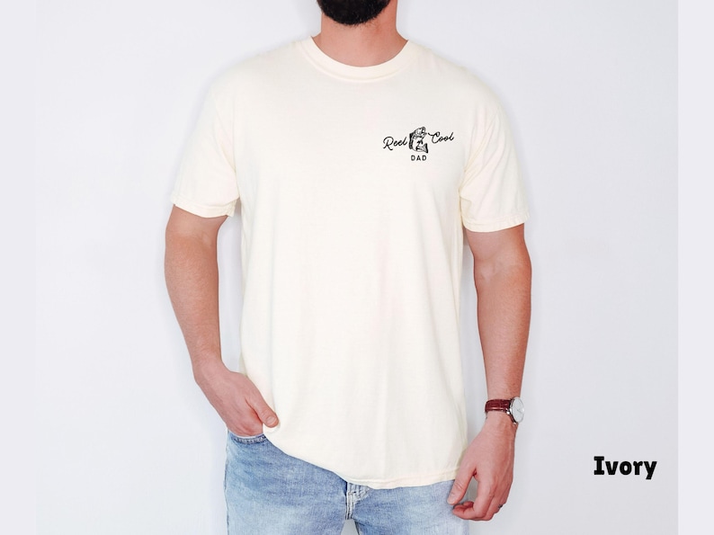 Pu&ograve; includere: T-shirt color avorio con la scritta "Reel Cool Dad" in corsivo nero. La maglietta ha girocollo e maniche corte. Il modello indossa jeans blu e un orologio con cinturino in pelle marrone.