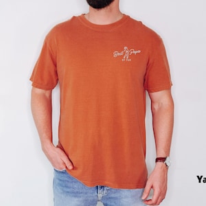 Op de afbeelding: Een oranje T-shirt met korte mouwen met de tekst "Best Papa" in witte letters. Het shirt wordt gedragen door een persoon die een spijkerbroek en een polshorloge draagt.