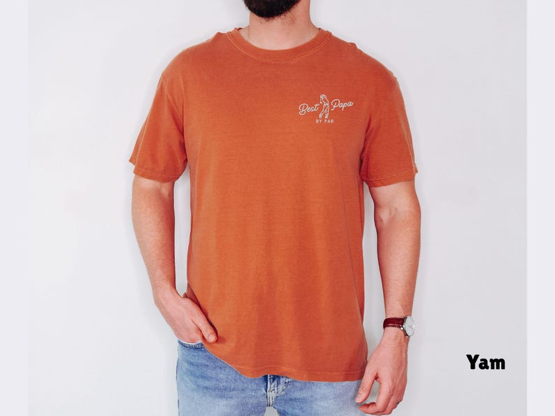 Op de afbeelding: Een oranje T-shirt met korte mouwen met de tekst "Best Papa" in witte letters. Het shirt wordt gedragen door een persoon die een spijkerbroek en een polshorloge draagt.