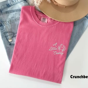 May include: A pink t-shirt with the text "A Little Crabby" and a crab graphic. The shirt is folded on top of a pair of light blue jeans and a tan hat. The shirt is labeled "Crunchberry".