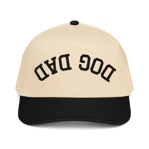 May include: A baseball cap with a cream-colored crown and a black brim. The text "DOG DAD" is embroidered in black, arched across the front. The cap has a black button on top.