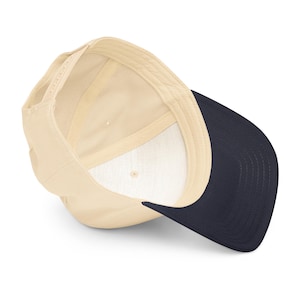 May include: A baseball cap with a cream-coloured crown and a navy blue brim. The interior of the cap is cream-coloured with a white sweatband. The cap has an adjustable snapback closure.