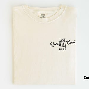 Op de afbeelding: Ivoor kleurig T-shirt met een ronde hals. Het shirt heeft een zwarte afbeelding van een vis en de woorden "Reel Cool Papa" in een scriptlettertype. Het T-shirt is opgevouwen en toont het ontwerp aan de voorkant.