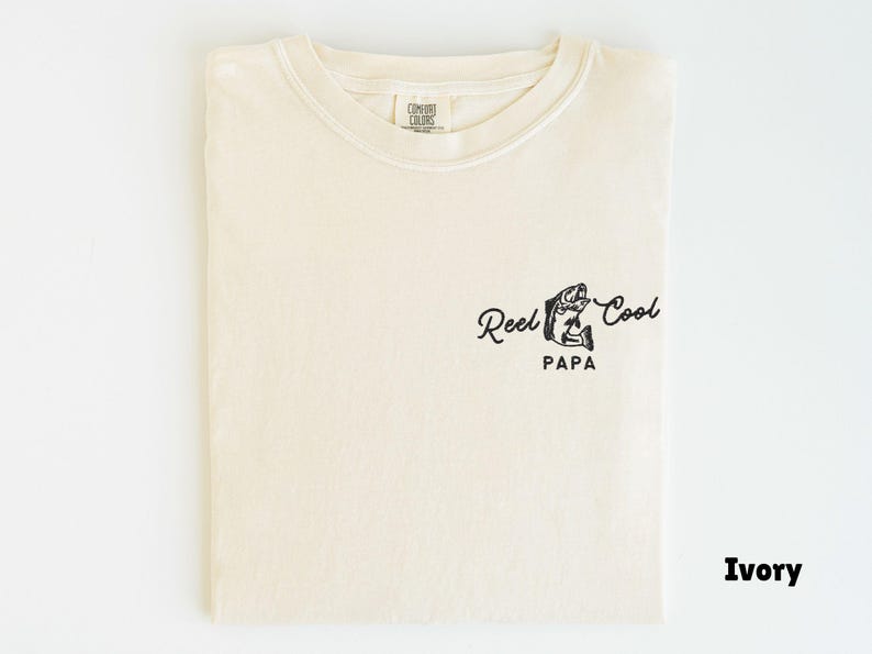 Op de afbeelding: Ivoor kleurig T-shirt met een ronde hals. Het shirt heeft een zwarte afbeelding van een vis en de woorden "Reel Cool Papa" in een scriptlettertype. Het T-shirt is opgevouwen en toont het ontwerp aan de voorkant.