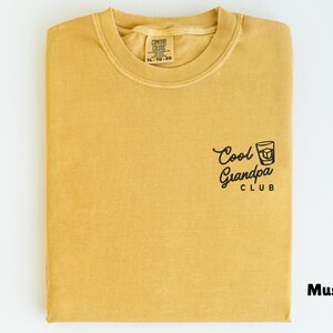 May include: Mustard-colored t-shirt with the text "Cool Grandpa Club" and a graphic of a drink. The shirt is folded and has a tag in the collar area. The shirt is a solid color.