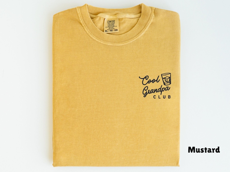 May include: Mustard-colored t-shirt with the text "Cool Grandpa Club" and a graphic of a drink. The shirt is folded and has a tag in the collar area. The shirt is a solid color.
