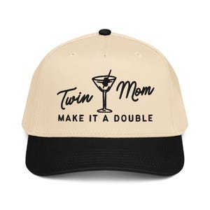 May include: A beige baseball cap with a black brim and a black button on top. The cap features the text "Twin Mom Make It A Double" in black script and a martini glass graphic.