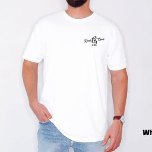 Pu&ograve; includere: T-shirt bianca con la scritta "Reel Cool Dad" in nero. La maglietta presenta una piccola grafica di un mulinello da pesca. Il modello indossa jeans blu e un orologio.
