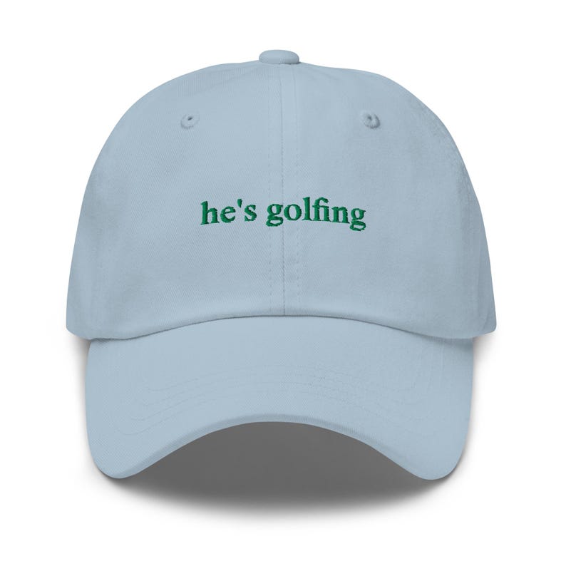 May include: A light blue baseball cap with the text "he's golfing" embroidered in green. The cap has a curved brim and a button on top. This hat is a stylish accessory for golf enthusiasts.
