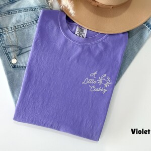 May include: A violet-colored t-shirt with the embroidered phrase "A Little Crabby" and a crab graphic. The shirt is folded and rests on a pair of blue jeans, with a tan hat and a gold necklace visible.