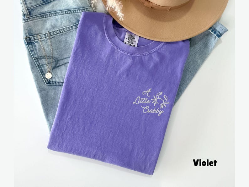 May include: A violet-colored t-shirt with the embroidered phrase "A Little Crabby" and a crab graphic. The shirt is folded and rests on a pair of blue jeans, with a tan hat and a gold necklace visible.