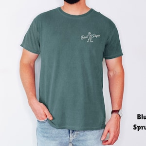 Op de afbeelding: Blauwspar T-shirt met de tekst "Best Papa BY FAR" in witte letters. Het shirt is effen van kleur en heeft een ronde hals. Het model draagt een blauwe spijkerbroek en een horloge.