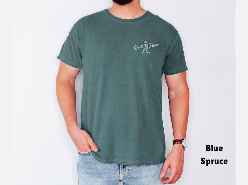 Op de afbeelding: Blauwspar T-shirt met de tekst "Best Papa BY FAR" in witte letters. Het shirt is effen van kleur en heeft een ronde hals. Het model draagt een blauwe spijkerbroek en een horloge.