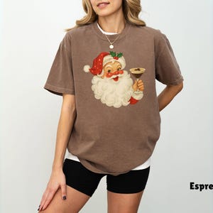 May include: Espresso-colored t-shirt featuring a vintage Santa Claus illustration holding a cocktail. Santa is wearing a red hat and suit with a white beard. The shirt is a casual style, perfect for the holiday season.