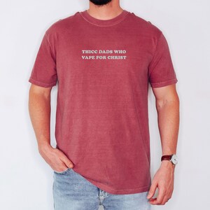 Puede incluir: Una camiseta granate con el texto "THICC DADS WHO VAPE FOR CHRIST" en gris claro. La camiseta la lleva una persona con vaqueros azules y un reloj.