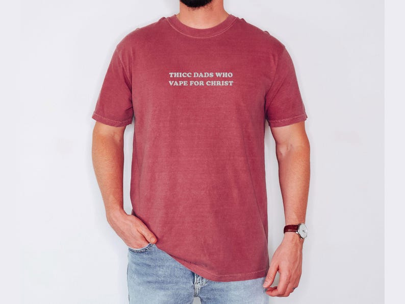 Puede incluir: Una camiseta granate con el texto "THICC DADS WHO VAPE FOR CHRIST" en gris claro. La camiseta la lleva una persona con vaqueros azules y un reloj.
