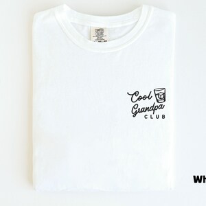 May include: White t-shirt with the text "Cool Grandpa Club" and a graphic of a drink. The shirt is folded neatly and has a tag at the neckline. The shirt is made of a soft material.