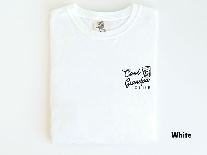 May include: White t-shirt with the text "Cool Grandpa Club" and a graphic of a drink. The shirt is folded neatly and has a tag at the neckline. The shirt is made of a soft material.