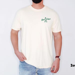 Op de afbeelding: Ivoor kleurig t-shirt met de tekst "Best Papa by Par" in het groen. Het shirt heeft een ronde hals en korte mouwen. Het model draagt een blauwe jeans en een horloge.