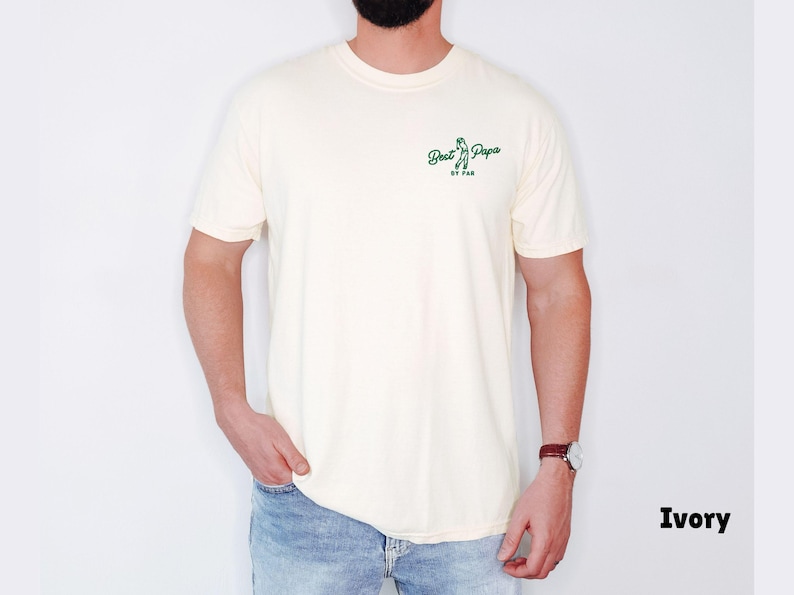 Op de afbeelding: Ivoor kleurig t-shirt met de tekst "Best Papa by Par" in het groen. Het shirt heeft een ronde hals en korte mouwen. Het model draagt een blauwe jeans en een horloge.