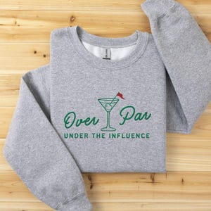 May include: A heather gray sweatshirt with the words "Over Par Under the Influence" in green script. A martini glass with a golf flag is in the center of the design. The sweatshirt is folded on a wooden surface.