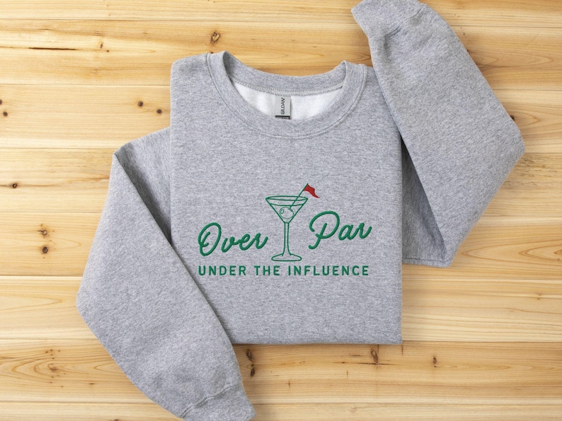 May include: A heather gray sweatshirt with the words "Over Par Under the Influence" in green script. A martini glass with a golf flag is in the center of the design. The sweatshirt is folded on a wooden surface.