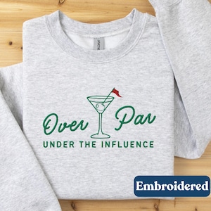 May include: A light gray sweatshirt with the words "Over Par Under The Influence" in green script, along with a martini glass graphic. The word "Embroidered" is in a blue box at the bottom.