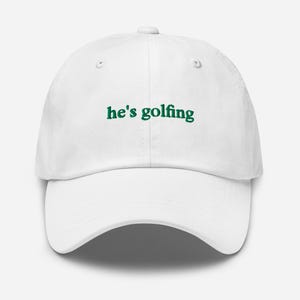 May include: White baseball cap with the text "he's golfing" in green lettering. The cap has a curved brim and a button on top. The hat is a classic style, suitable for outdoor activities.