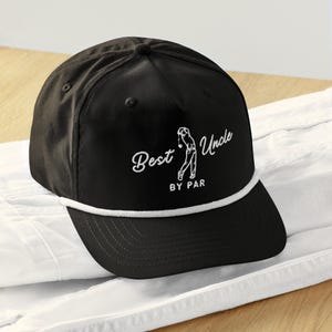 May include: Black golf cap with white embroidered text that reads "Best Uncle By Par" and a golf player graphic. The cap has a white rope detail along the brim and a curved visor. The cap is sitting on a white surface.