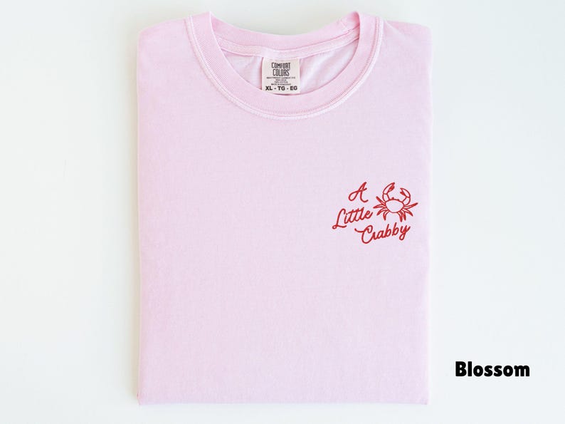 May include: A light pink t-shirt with the words "A Little Crabby" and a crab graphic embroidered in red. The shirt is folded and has a tag that reads "Comfort Colors" and the size "XL - TG - EG".