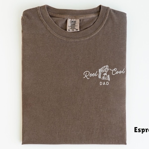 Pu&ograve; includere: T-shirt color espresso con la scritta ricamata "Reel Cool Dad" e una grafica di pesce. La maglietta &egrave; piegata, mostrando il disegno sul davanti. L'etichetta indica XL, e la maglietta &egrave; in materiale morbido.