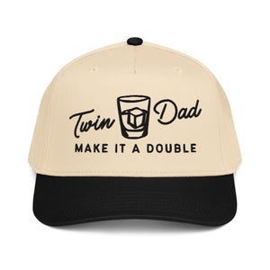 May include: A beige baseball cap with a black brim. The cap features the text "Twin Dad MAKE IT A DOUBLE" in black script and block letters, along with a graphic of a drink with ice cubes.