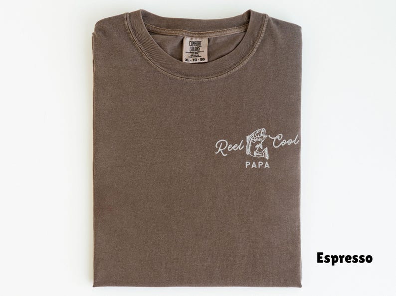 Op de afbeelding: Espresso-kleurig T-shirt met de witte tekst "Reel Cool Papa" en een afbeelding van een vis en hengel. Het gevouwen shirt heeft een label in het midden bovenaan.
