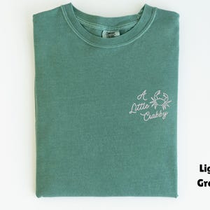 May include: Light green t-shirt with a crew neck. The shirt has the embroidered phrase "A Little Crabby" with a crab graphic on the front. The shirt is folded neatly on a white surface.